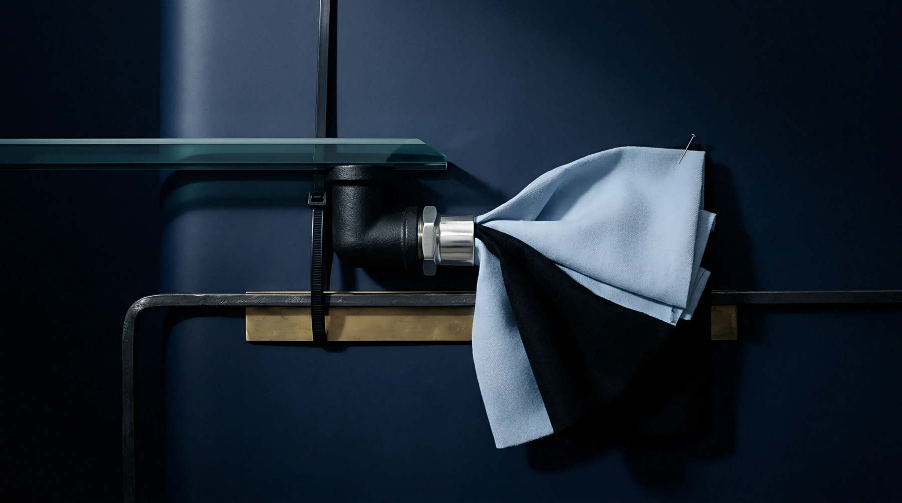 A folded light blue and black fabric swatch is pinned to a silver collar connecting matte black industrial pipes against a deep navy wall.