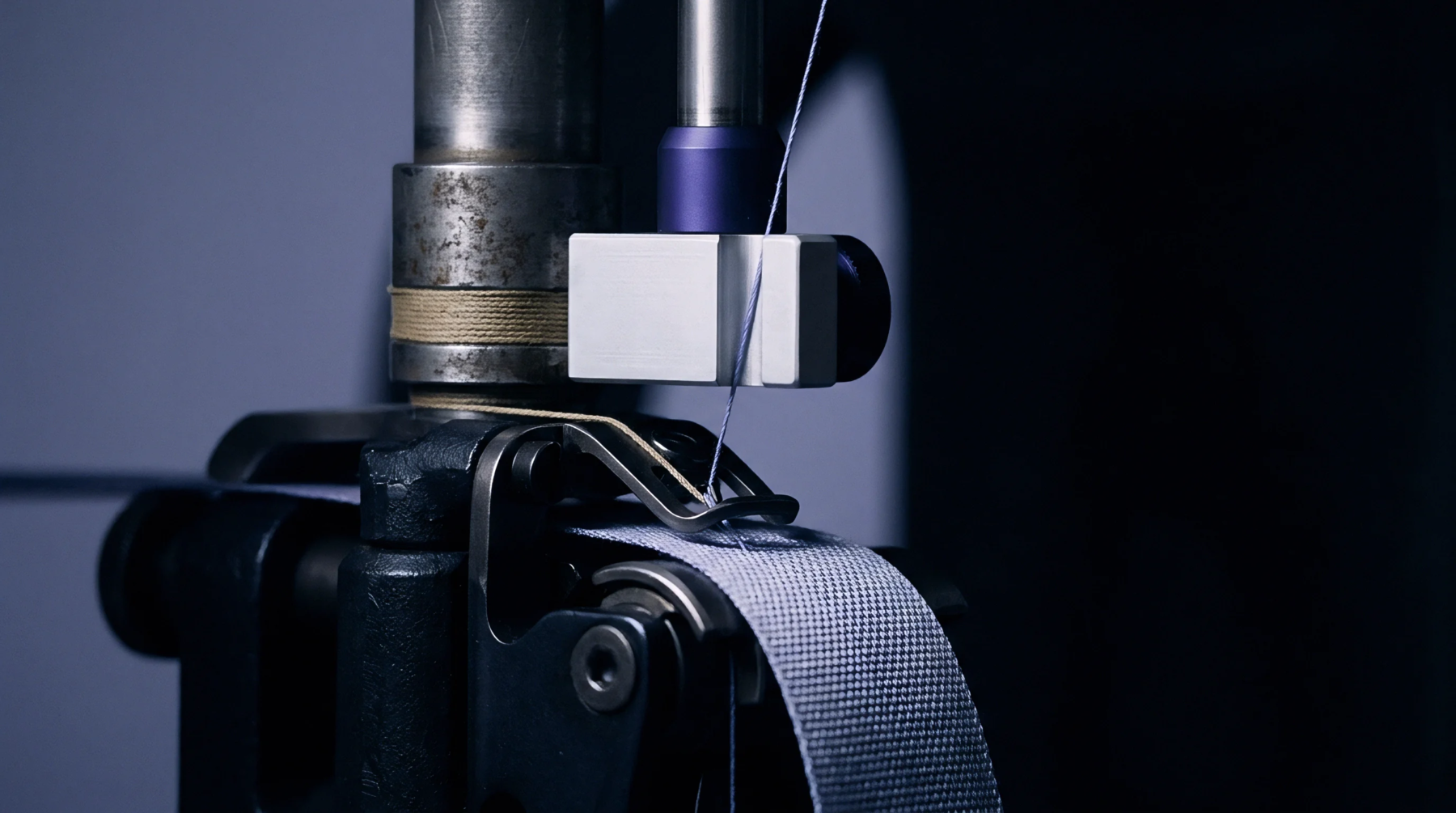 A metal guide feeds blue thread into a thick grey woven textile strap positioned on the needle bed of industrial machinery.