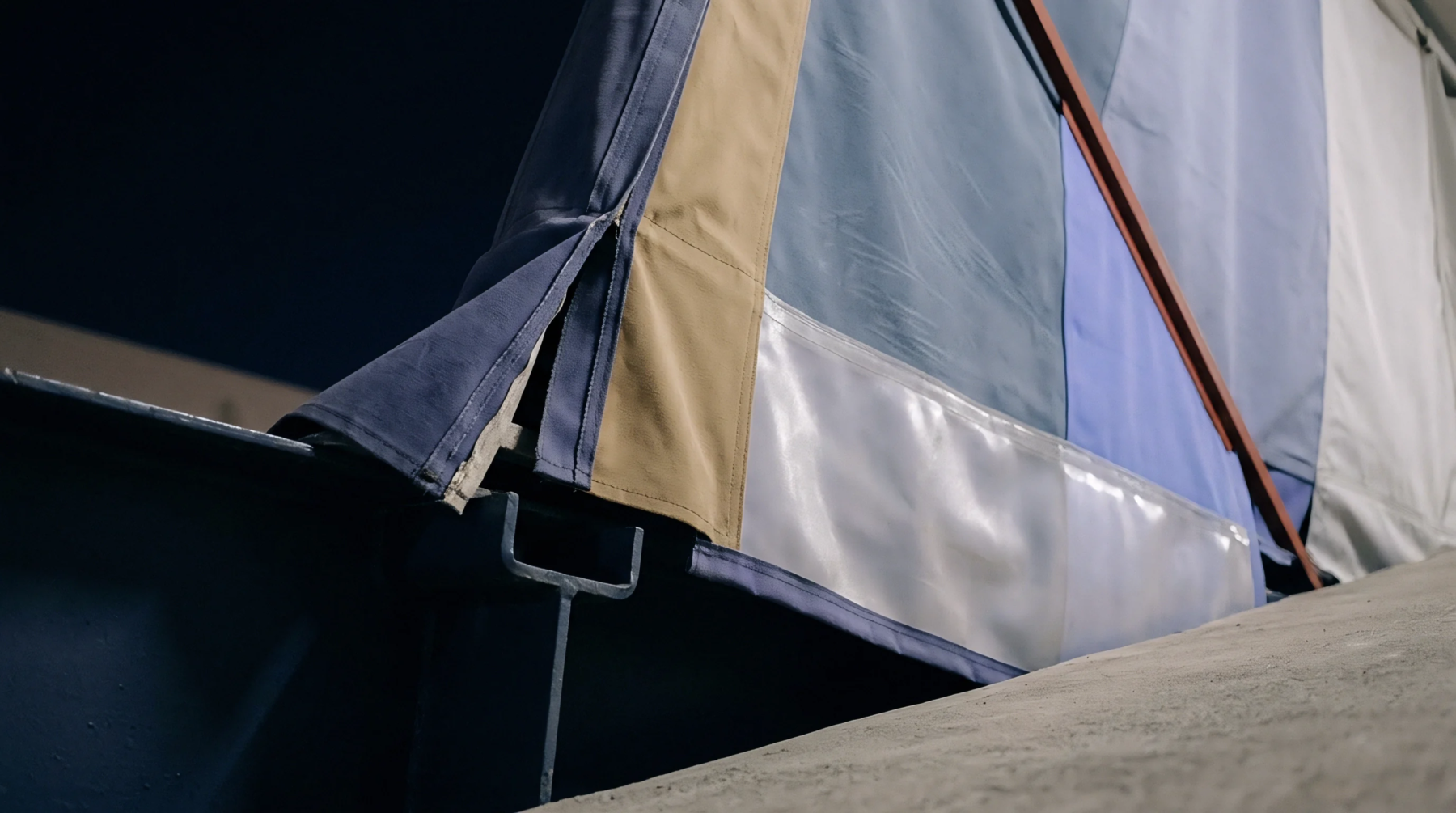 A multi panel industrial fabric curtain constructed from tan canvas, blue synthetic sheets, and a clear vinyl window strip hangs above a concrete floor.