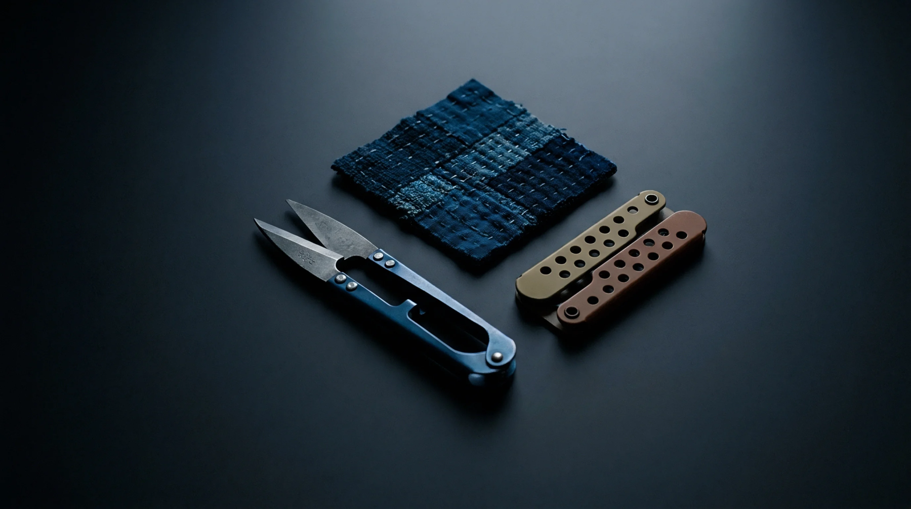 Precision thread snips and folding hardware rest beside a dark indigo dyed sashiko stitched textile patch on a matte surface.