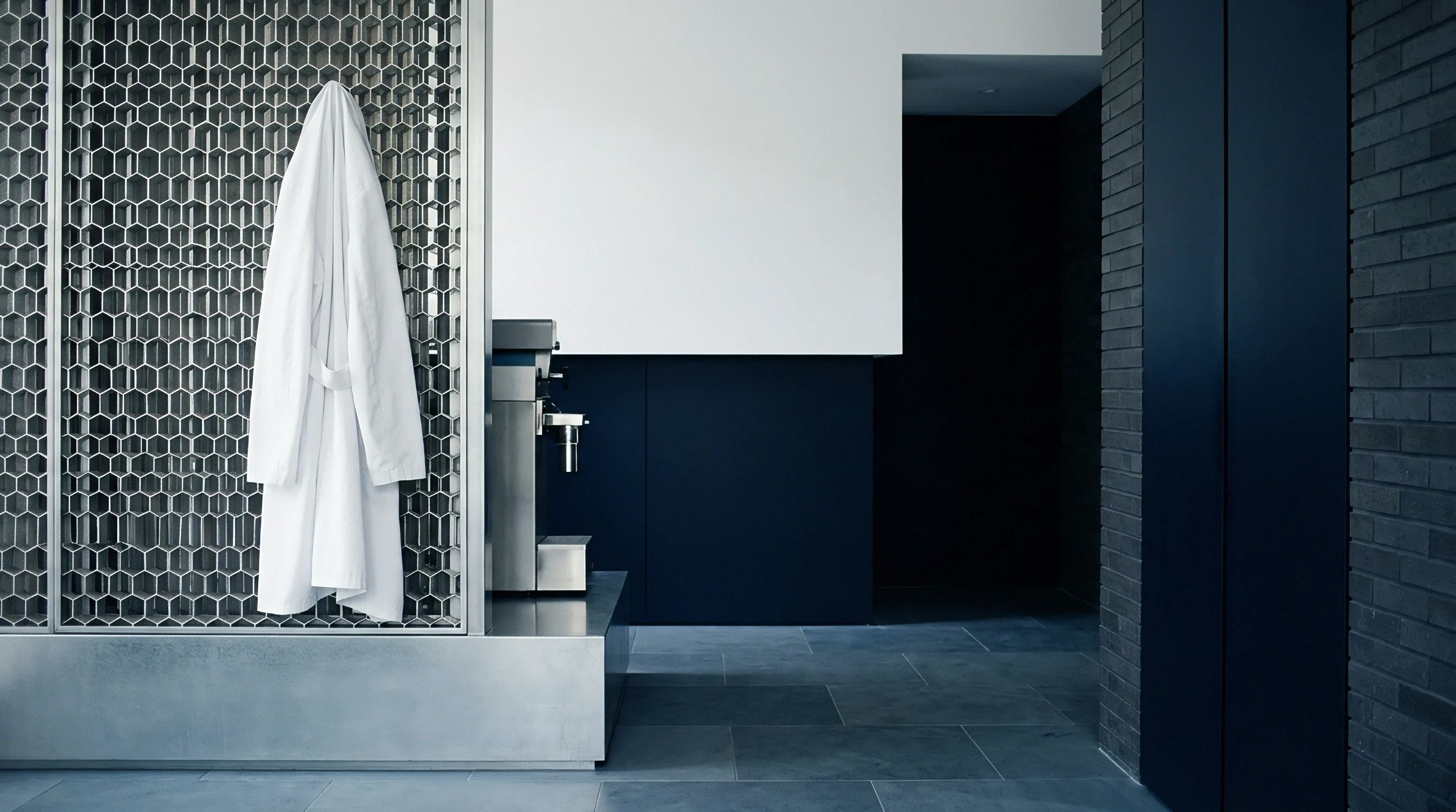White terry cotton bathrobe hangs against a silver hexagonal metal lattice structure positioned within a minimalist industrial interior setting featuring dark slate flooring.